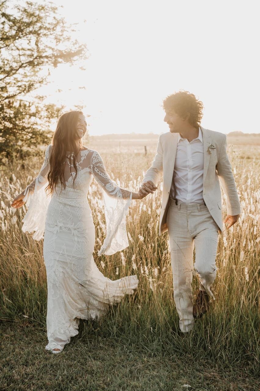 Novios caminando de la mano entre pastizales dorados en su casamiento